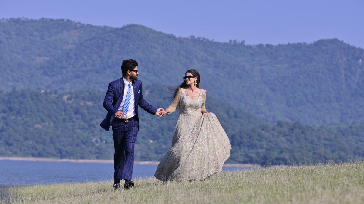 A couple in elegant wedding attire walking hand in hand by a picturesque riverbank in Una, India.